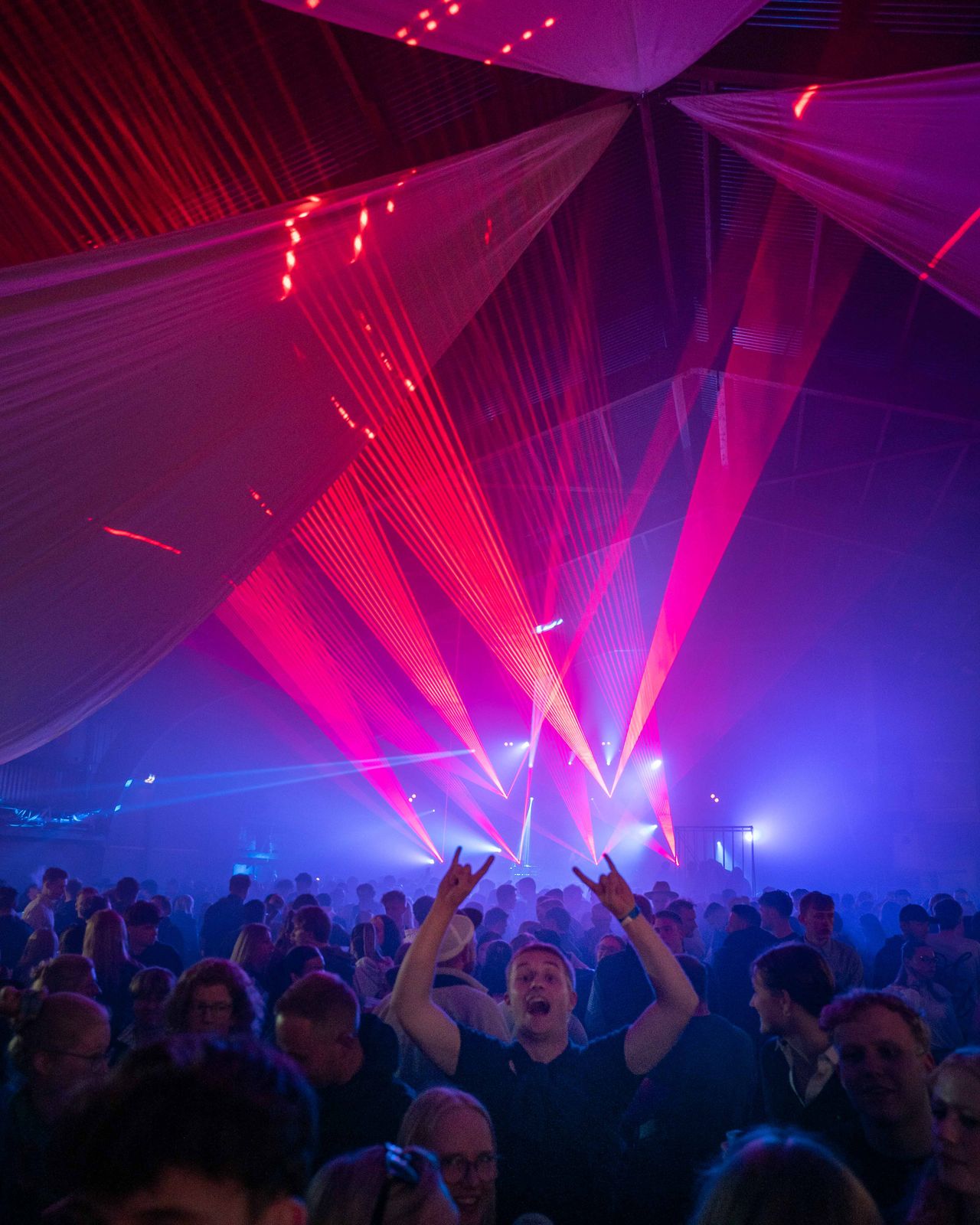 Blaues Licht und rote Laser von der Bühne. Ein Gast reißt Hände in der Crowd hoch. – Maifeuerfete Fehmarn 2026