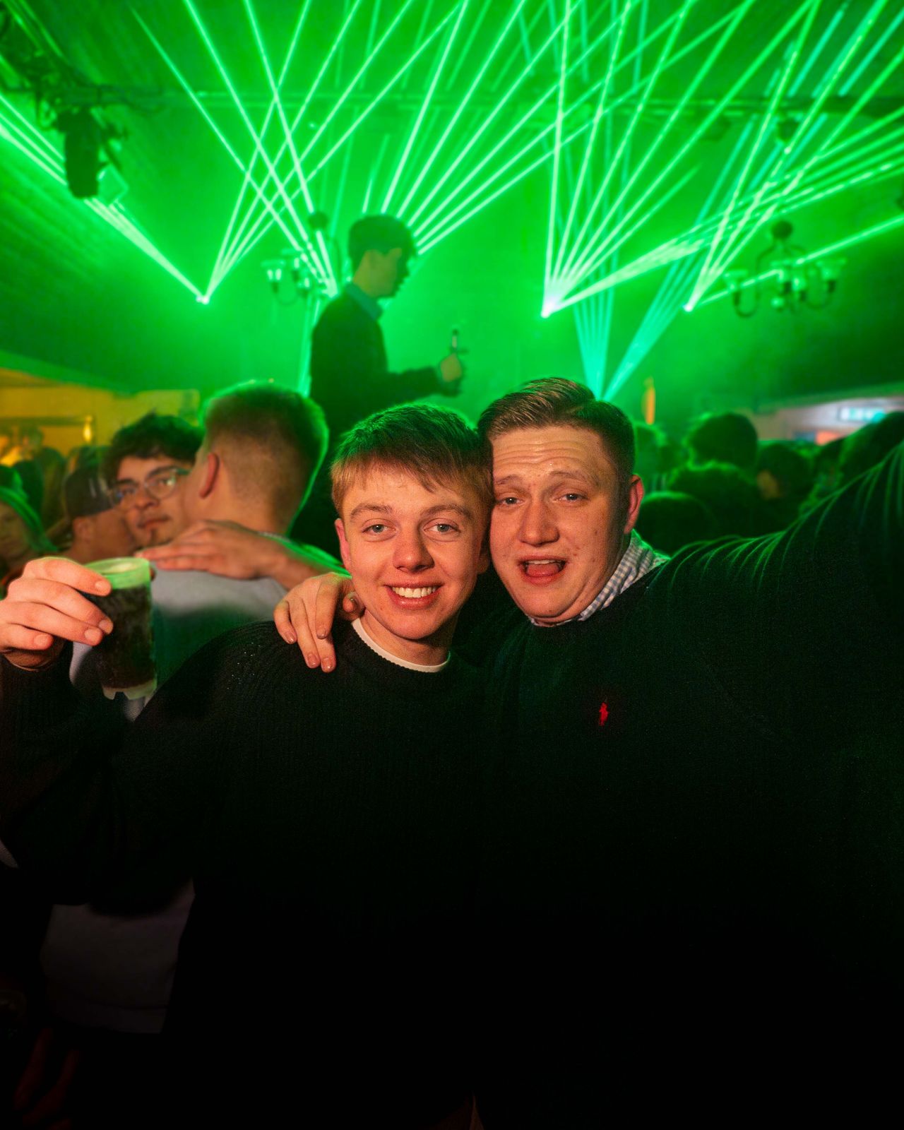 Zwei Gäste Arm in Arm gucken in die Kamera. Im Hintergrund sind grüne Laserstrahlen zu sehen.