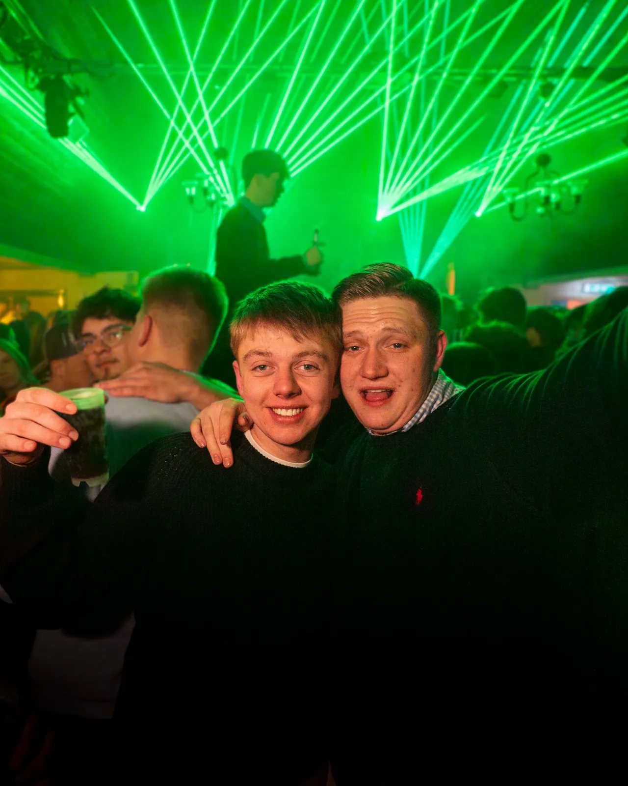 Zwei Gäste Arm in Arm gucken in die Kamera. Im Hintergrund sind grüne Laserstrahlen zu sehen.