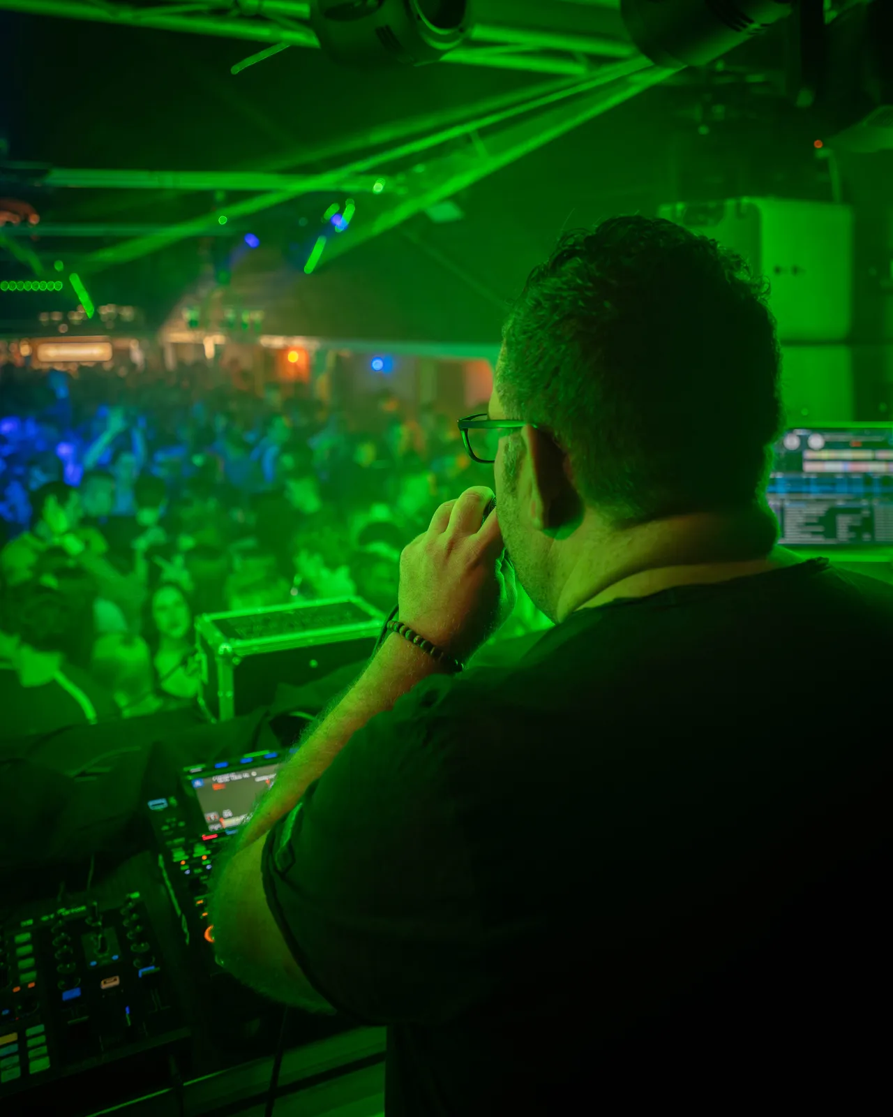 Basti van den Sroeden mit Mikrofon am DJ Pult im grünen Licht. Im Hintergrund ist die Crowd zu sehen.