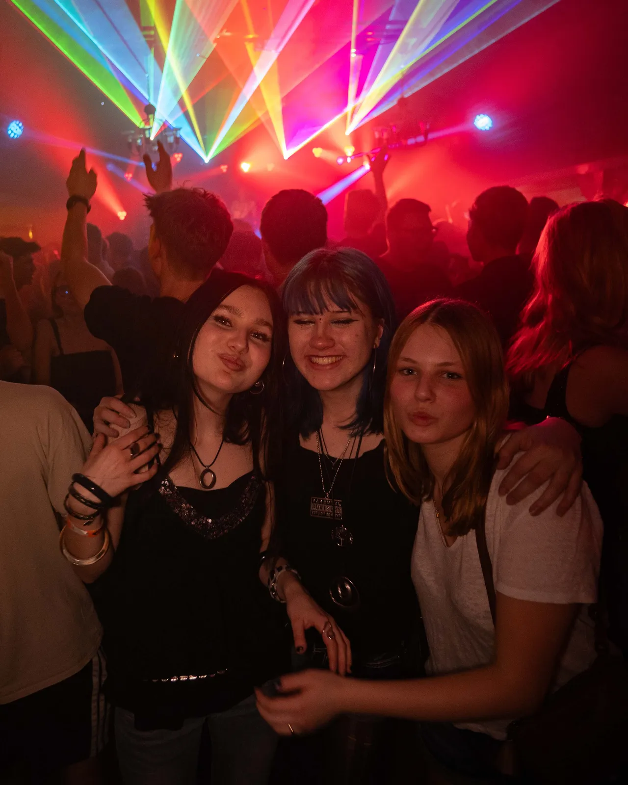 Drei Gäste schauen in die Kamera. Im Hintergrund ist roten Licht und bunte Laserstrahlen zu sehen.