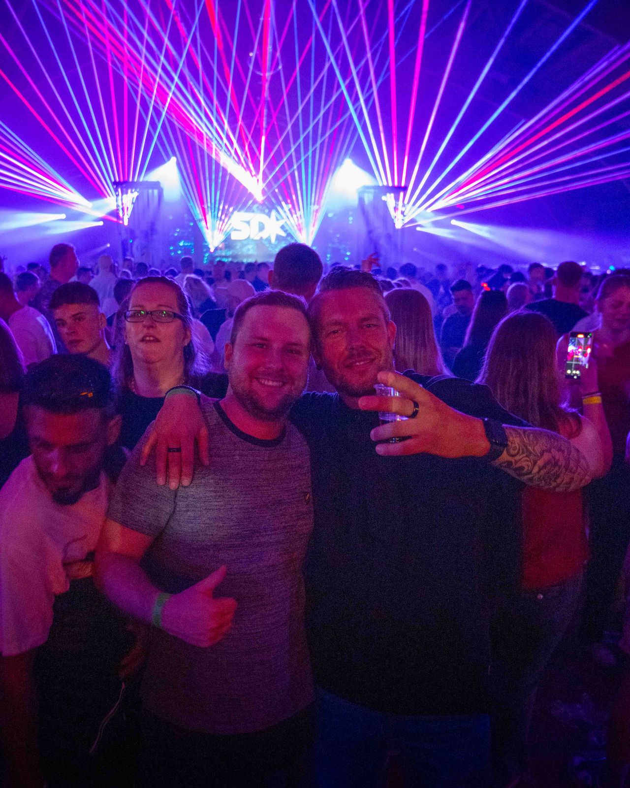 Zwei Gäste mit Rücken zur Bühne und Laser im Hintergrund gucken in Kamera.